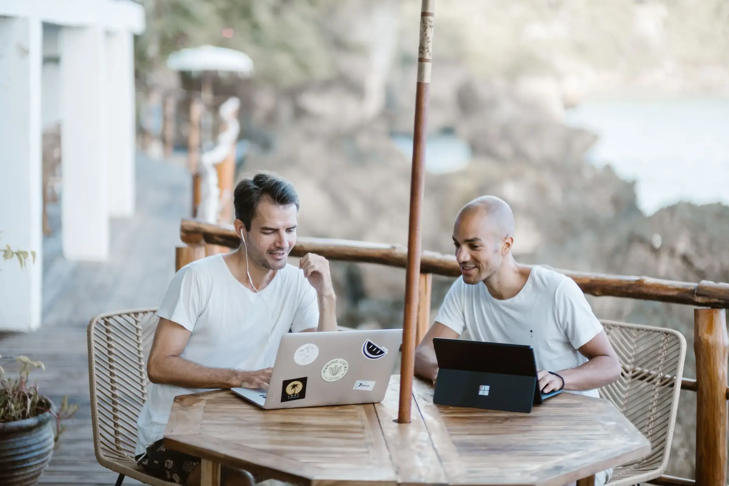 Zwei Männer arbeiten gemeinsam an Laptops an einem Holztisch im Freien